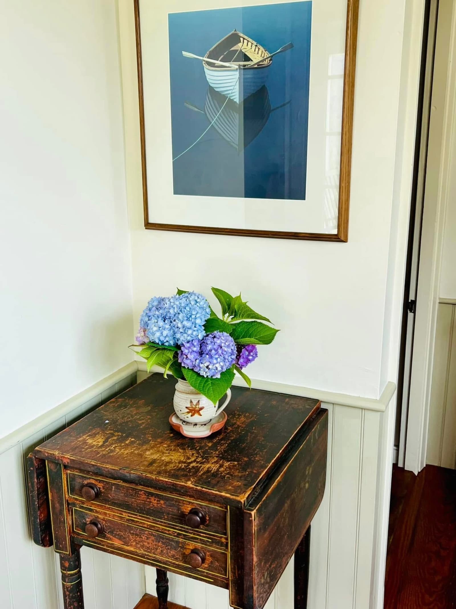 Antique entryway table with hydrangeas in pottery vase and rowboat painting above